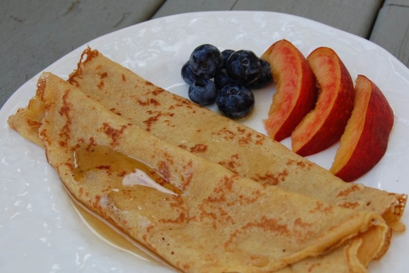 Two crepes on a plate with peach slices and blueberries. 
