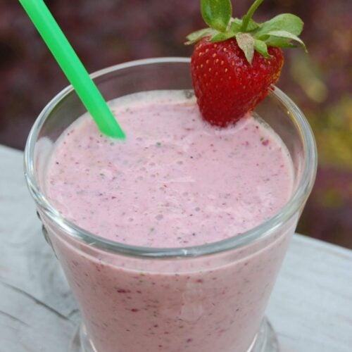 A strawberry smoothie in a glass with a fresh strawberry on the side and green straw.