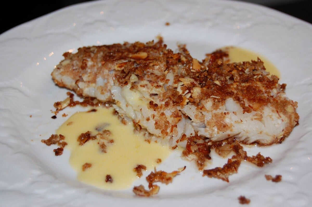 A piece of white fish almond encrusted drizzled with a Beurre Blanc Sauce on a white dish.