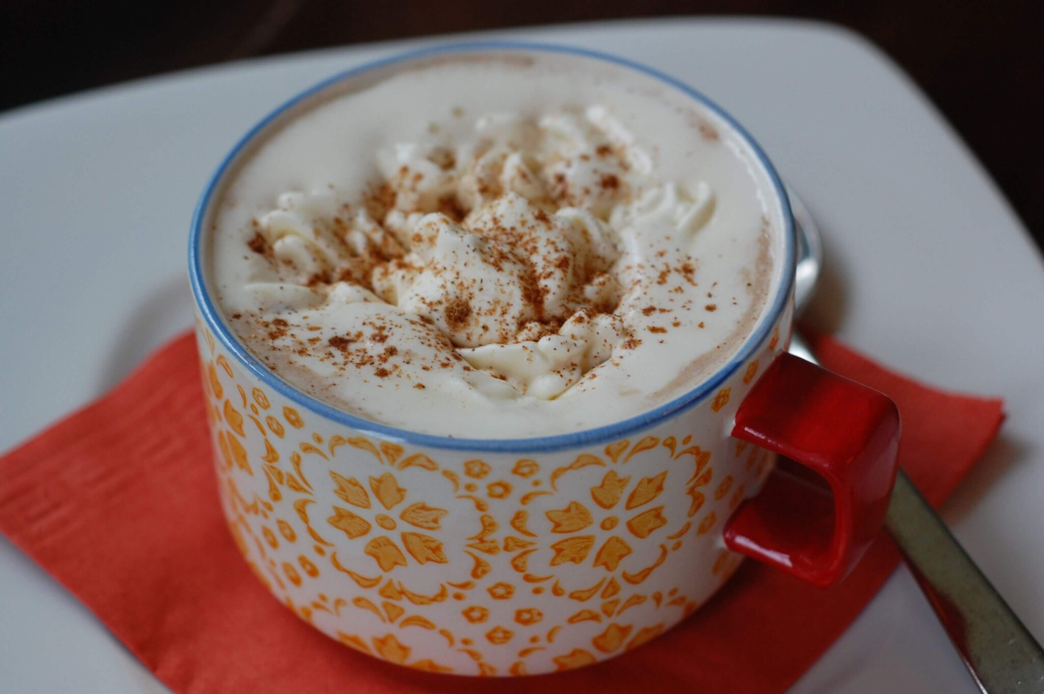 A decorative yellow and white mug with a red handle is filled with creamy cappuccino topped with whipped cream and a sprinkle of cinnamon. The mug sits on a red napkin on a white plate with a spoon placed beside it.