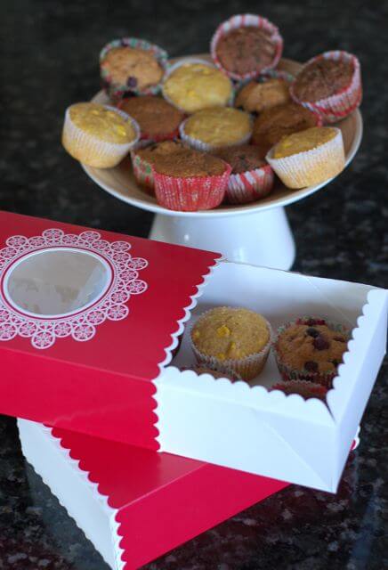 Assorted muffins in decorative muffins tins sitting on a white cake stand and in holiday boxes. 