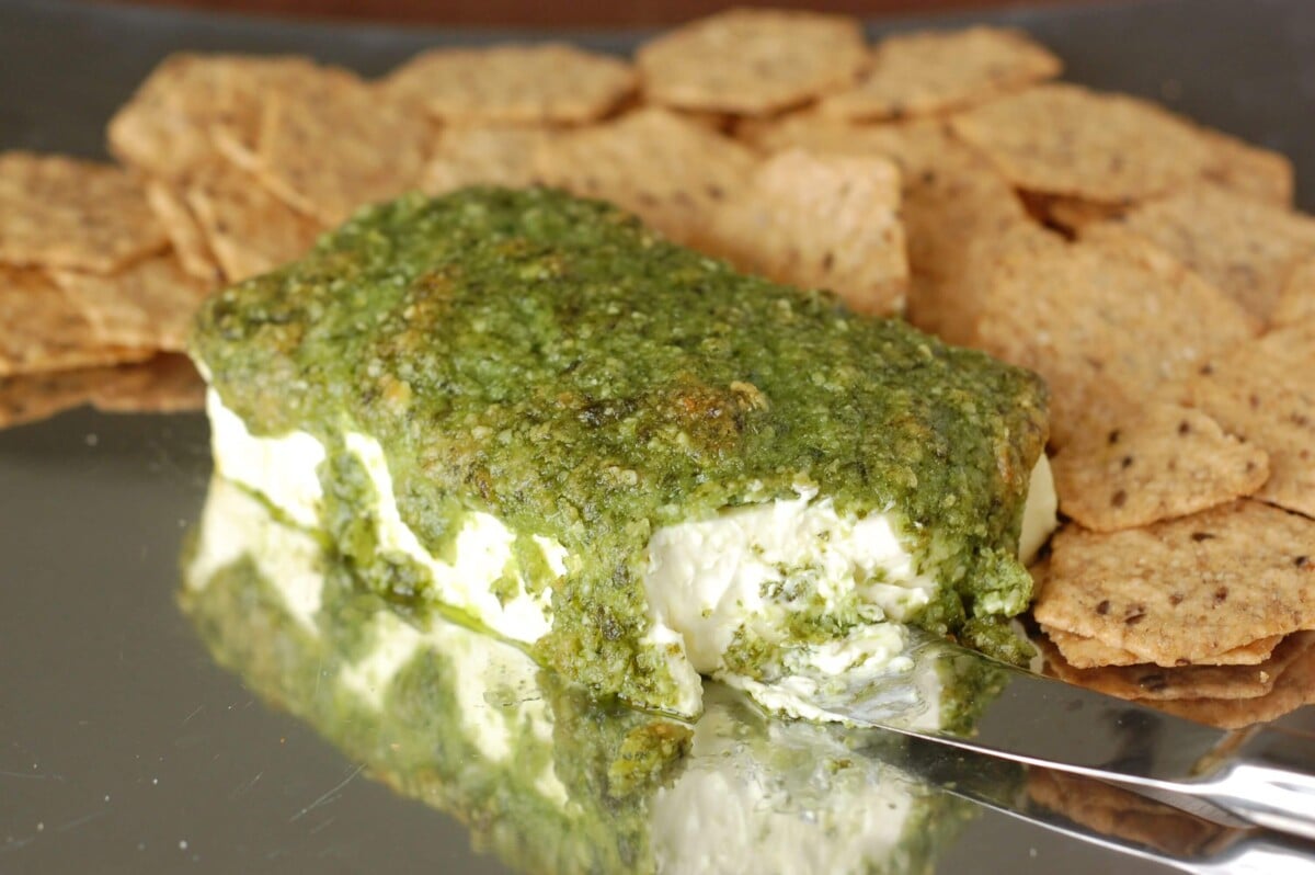 Block of cream cheese covered in pesto with whole-grain crackers around it.