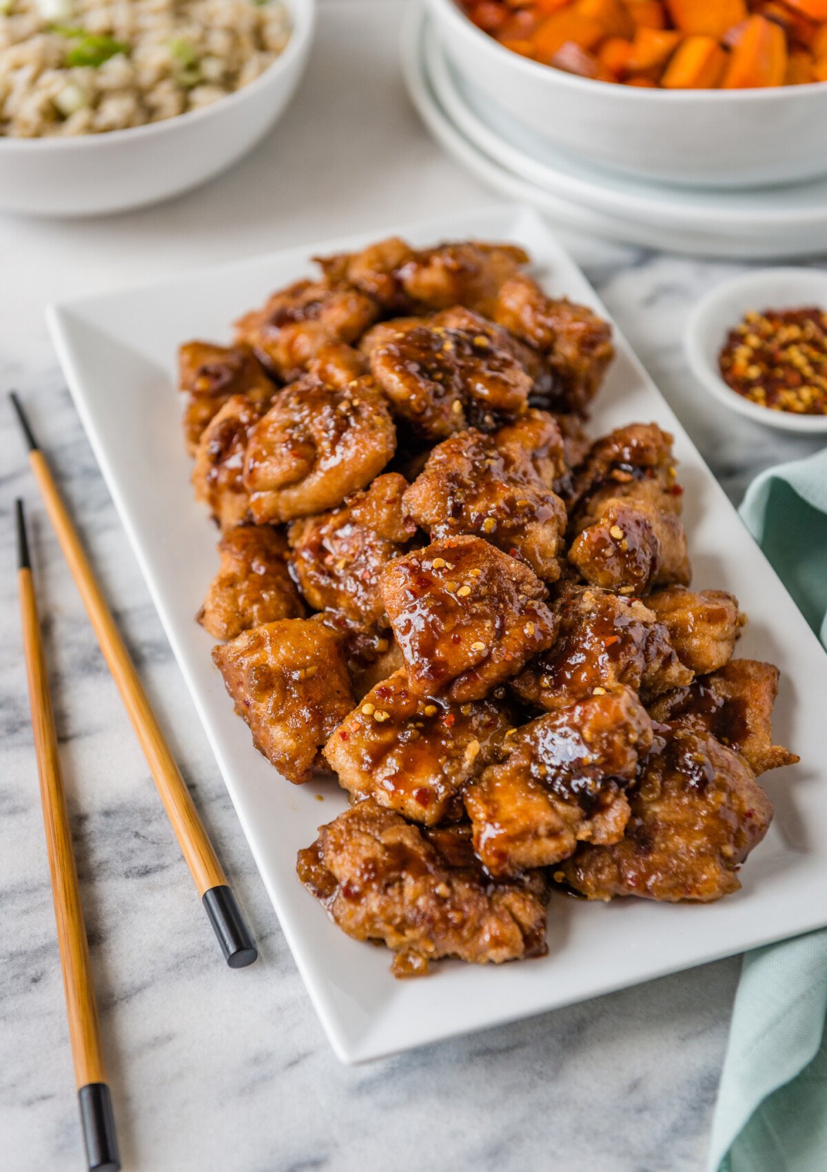 A serving platter of homemade Chinese chicken topped with red pepper flakes, along side different side dishes and chopsticks. 