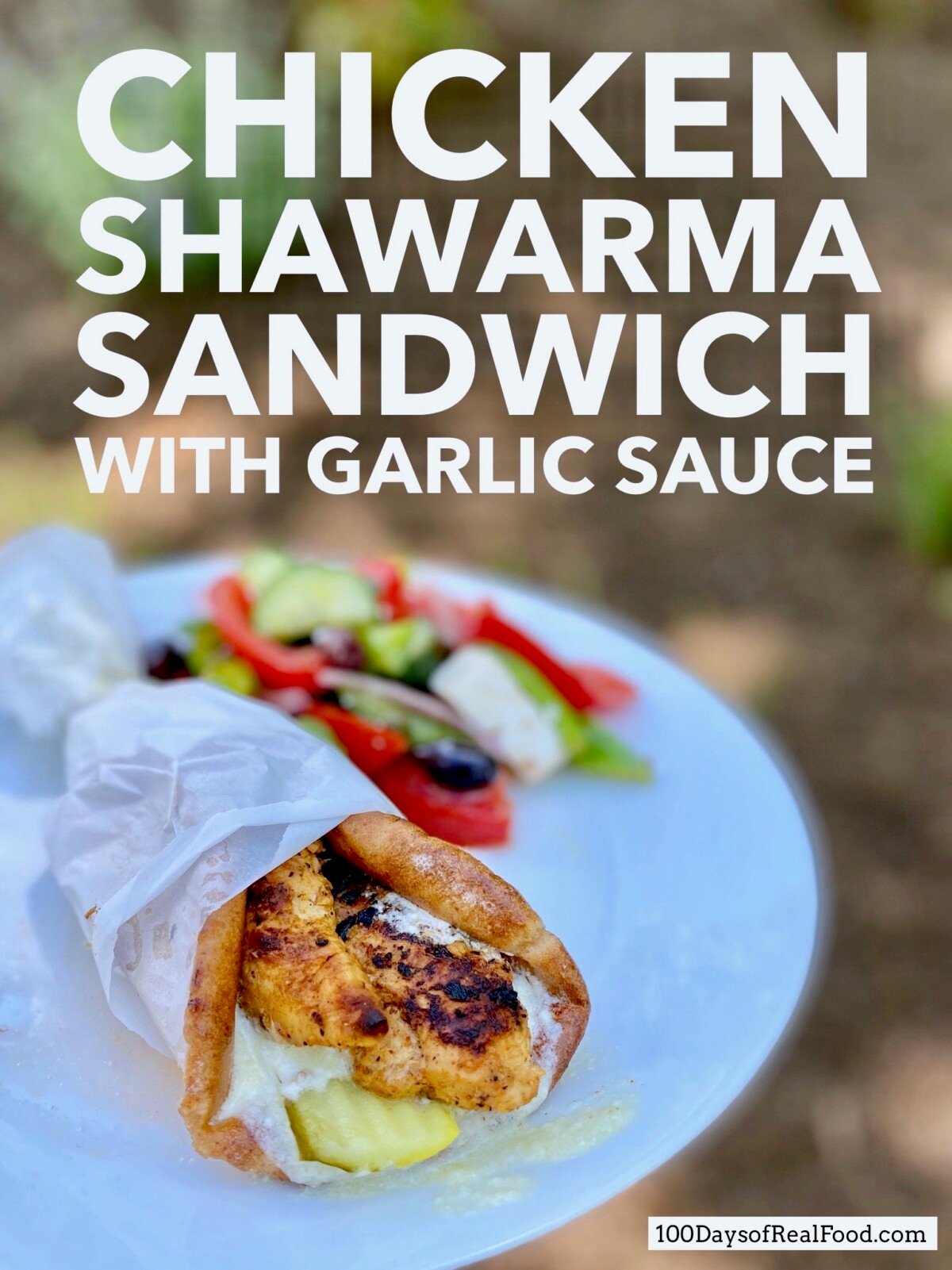 A Pita filled with grilled chicken, a pickle, and garlic sauce wrapped in parchment paper on a plate with a colorful side salad.