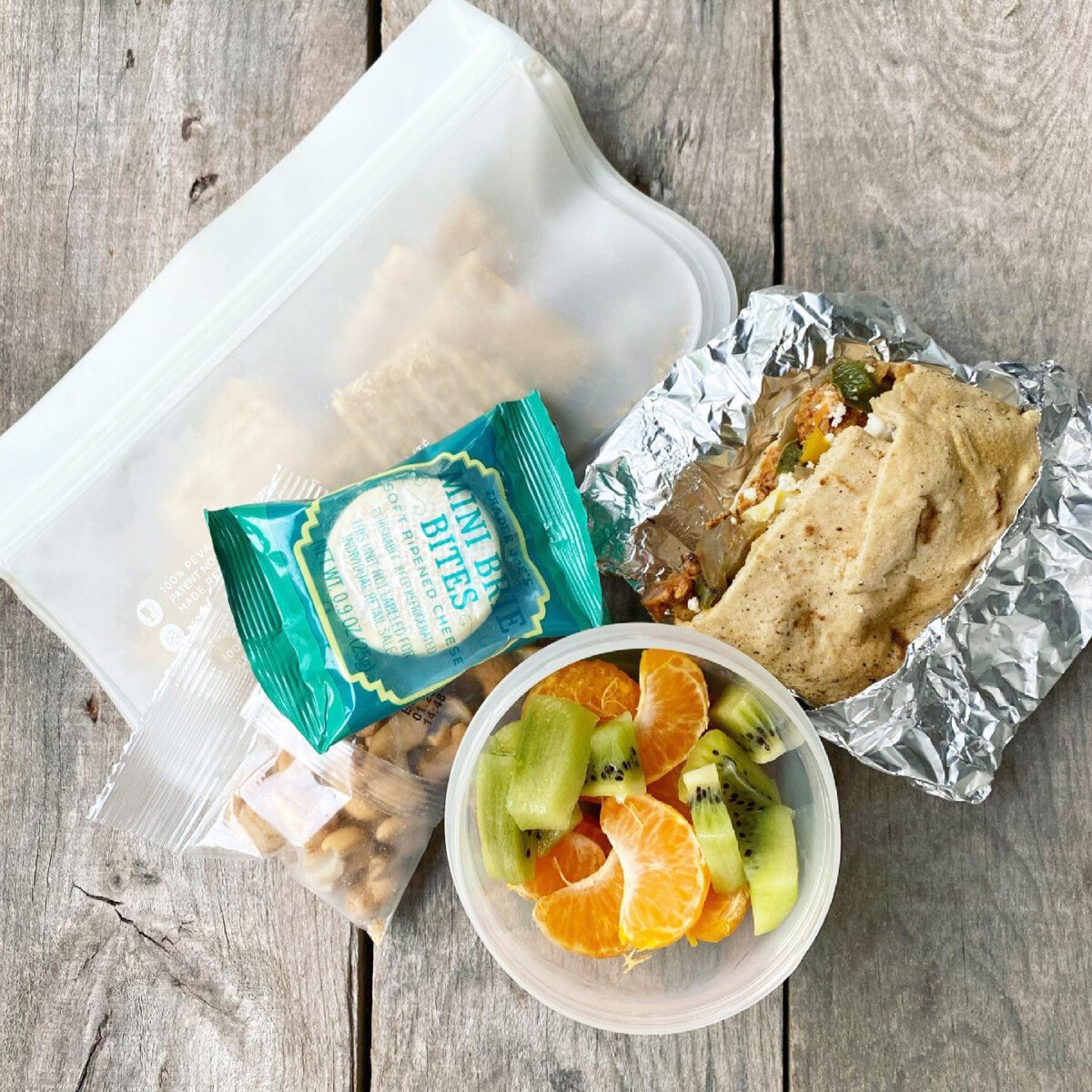 Packed school lunch that includes a Mexican Breakfast Burrito, kiwi and oranges, cashews, and whole-wheat crackers with a mini brie.