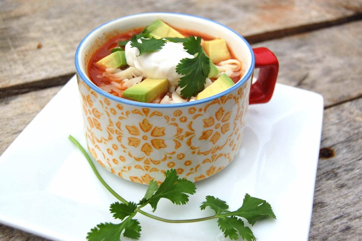 A colorful mug filled with Tortilla Soup topped with sour cream, avocado, and cilantro. 