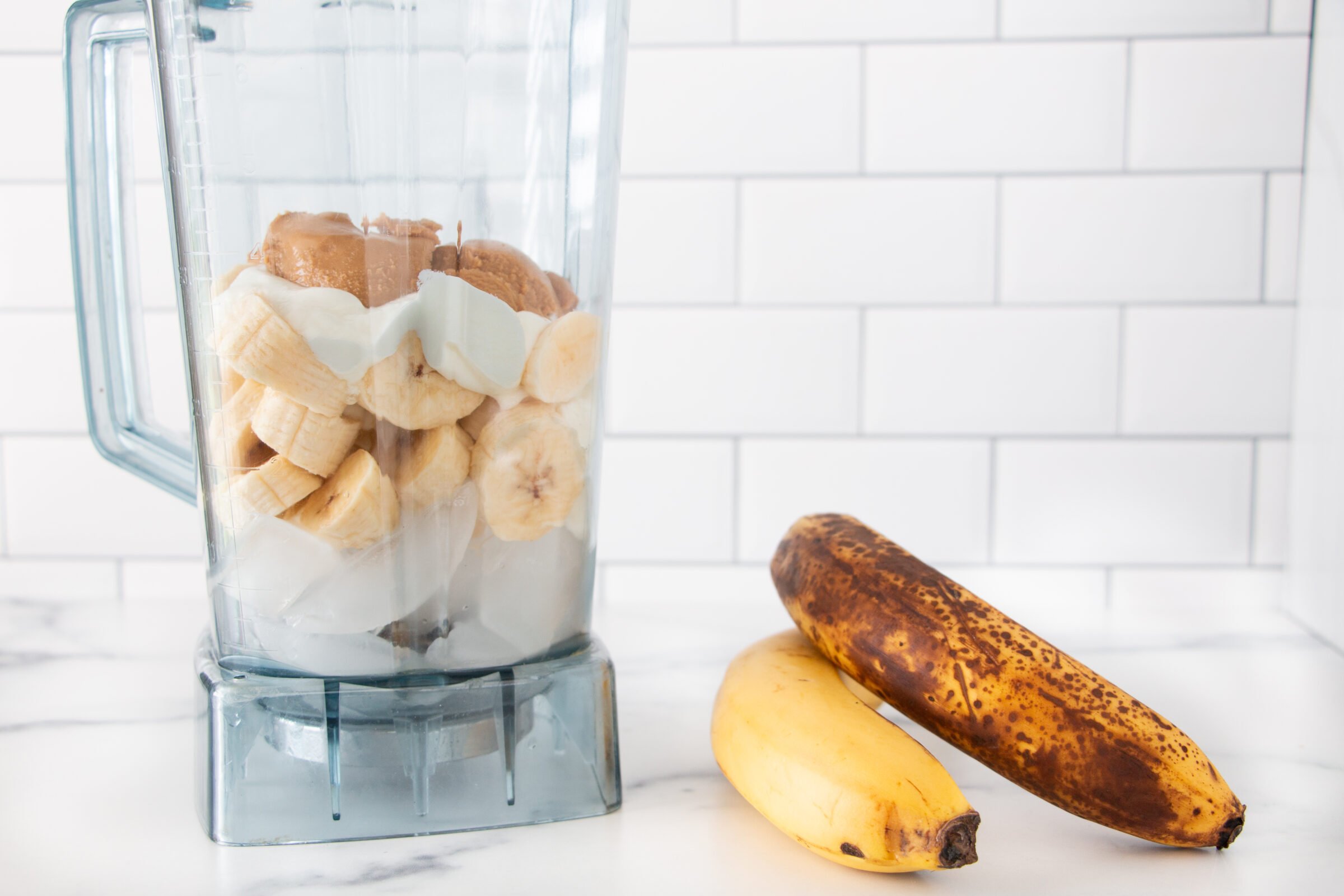 Blender with fresh ingredients of ice, banana slices, yogurt, and peanut butter layered to make a Peanut Butter Banana Smoothie.