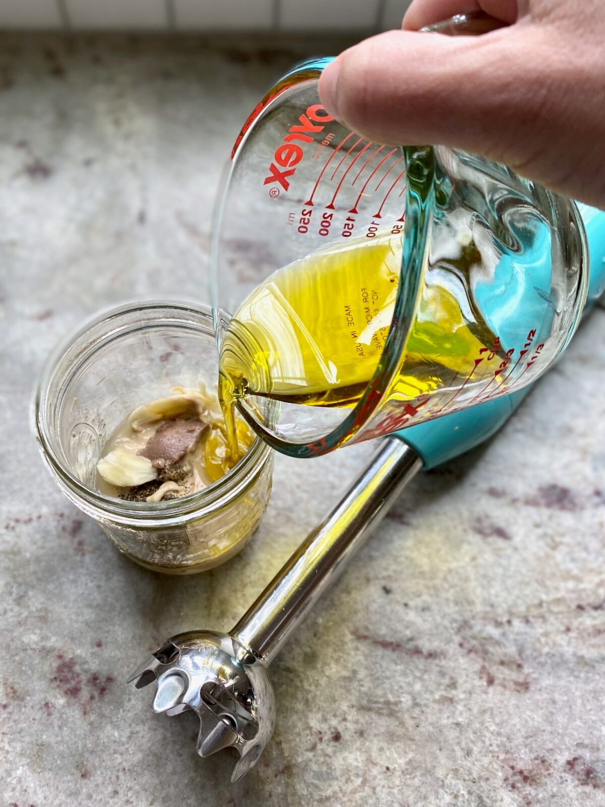 Pouring olive oil into a mason jar with remaining ingredients to make homemade Caesar dressing.