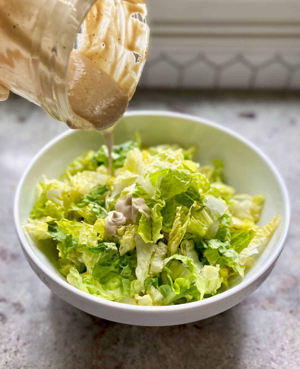 Pouring homemade Caesar dressing onto a bowl of chopped romaine lettuce to make Smoked Salmon Caesar.