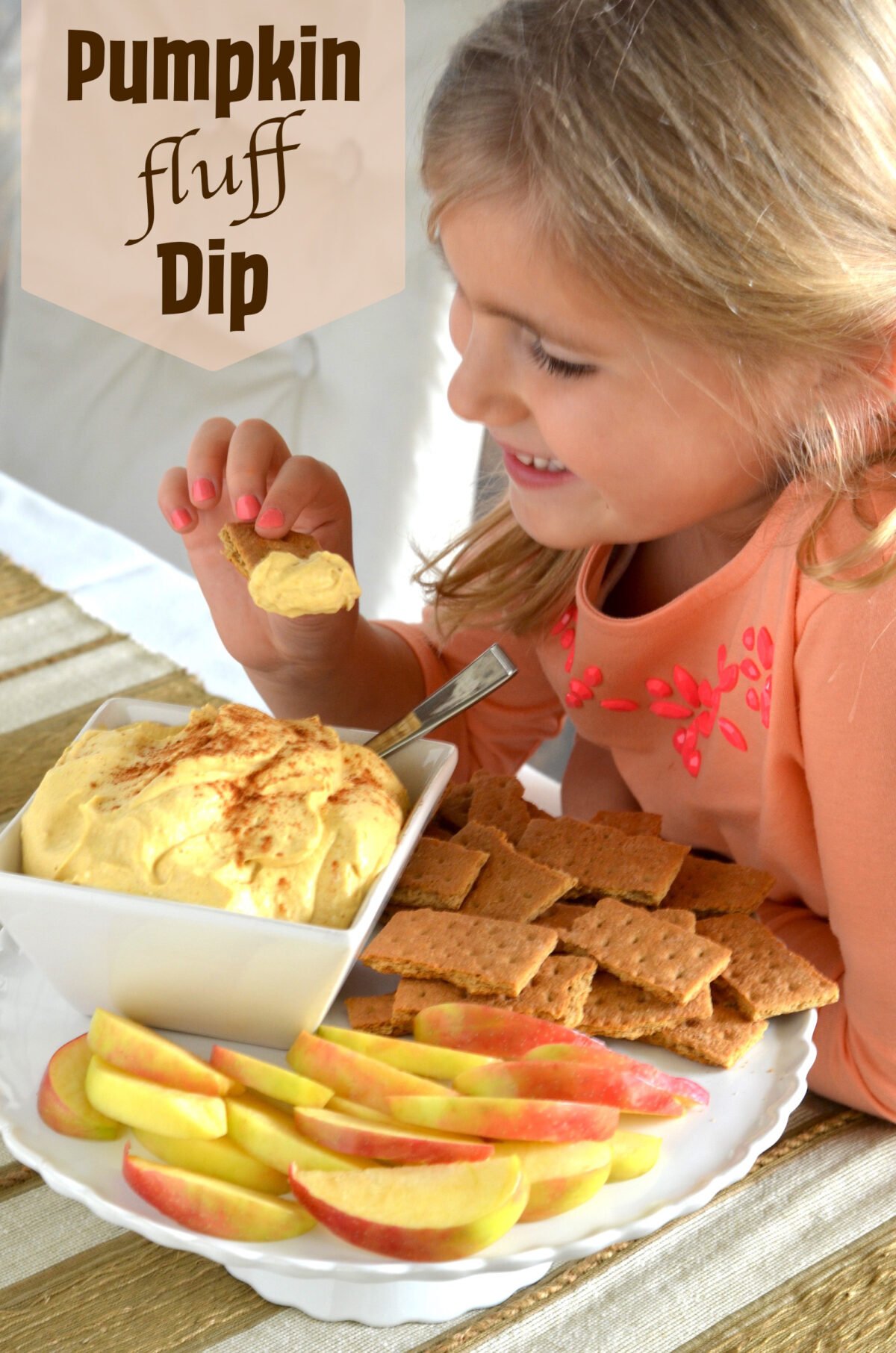 Young girl happily eating pumpkin fluff dip on graham crackers with a side of apple slices.